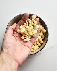 White chocolate drops in mans hand above on the bowl on white marble background. Baking ingredient