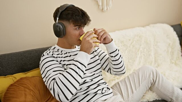 Confident young hispanic man chillin' on the sofa, headphones on, lost in music, sipping hot coffee in his cozy living room