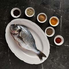 Raw and fresh dorado fish on black marble background with spices for cooking around. Minimalistic, dark-style or low key food photo. Mediterranean diet. Top view, copy space