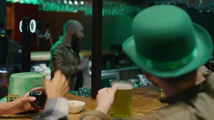 Over shoulder footage of unidentified man and woman in green hats calling Irish friends and congratulating on Saint Patricks day via video call while celebrating at bar counter with green beer in pubO