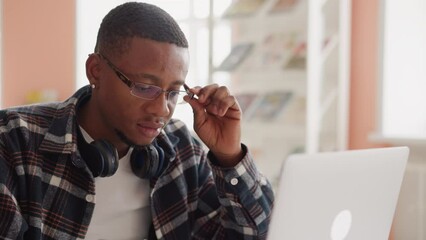 Student in glasses uses computer in library. African American man with earphones takes off glasses looking attentively at screen. Exams preparation at university