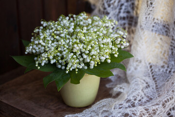 Bouquet of spring lily of the valley flowers in a vase on a wooden chair, white lace shawl, blur, still life.