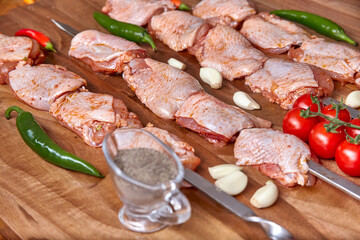 Metal skewers with raw uncooked chicken meat for frying on the wooden board with peppers near it, close-up perspective view. Shallow depth of field. Chicken, cherry tomato, pepper and garlic in focus