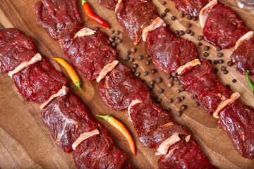 Metal skewers with raw uncooked beef meat for frying on the wooden board with chili peppers and pepper near it, close-up perspective view shallow depth of field. Meat, peppercorns and pepper in focus