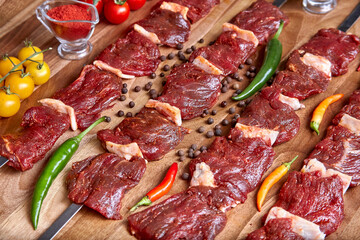 Metal skewers with raw uncooked beef meat for frying on the wooden board with peppers, spices and cherry tomatoes near it, close-up perspective view shallow depth of field. Meat and spices in focus