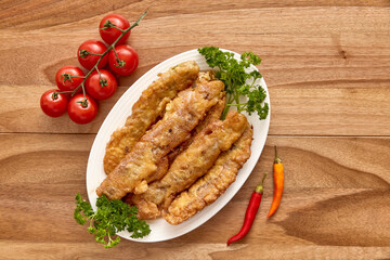 White plate with breaded meat on the wooden table with cherry tomatoes, chili pepper and greens near it. Top view, flat lay