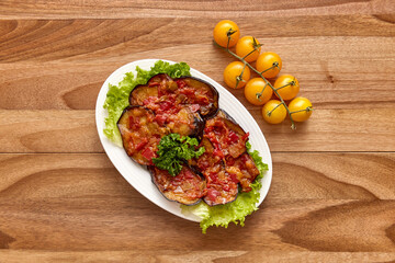 White plate with fried eggplant with tomato sauce on a lettuce leaf on the wooden table with cherry tomatoes near it. Top view, flat lay