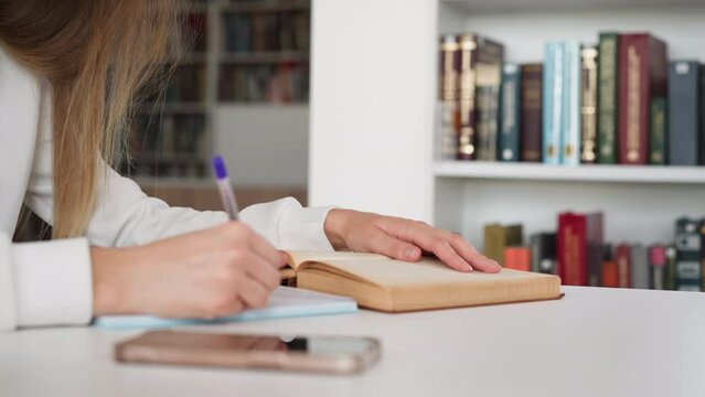 Female student rewrites information from book into notebook. Young woman prepares for seminar in university library. Obtaining higher education