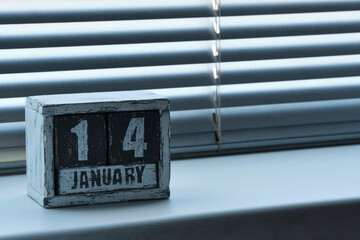 Morning January 14 on wooden calendar standing on window with blinds.