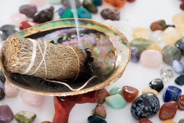 A close up image of a burning sage smudge stick with small healing crystals on a white table. 
