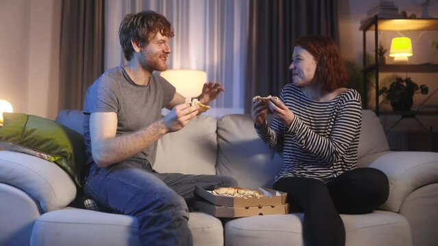 Smiling young couple eating pizza and talking sharing news after hard working day at cozy home Happy people enjoying spending time together sitting on couch at late evening indoors