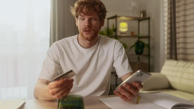 Confused red haired man with credit card thoughtfully pondering on screen while has problem with online order payment error or blocked bank account doing shopping at internet on smartphone at home
