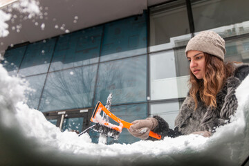 Young  woman using a snow brush to remove snow from her car's windshield. Concept of winter weather, car maintenance, and being prepared for snowy conditions. Transportation concept