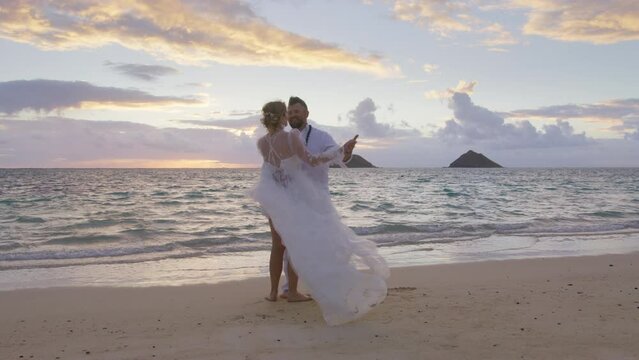 Romantic happy hugging young couple sharing beautiful first wedding dance moment on Hawaii beach at sunset. In love partners enjoying elopement beach ceremony. Carefree loving people dancing on beach