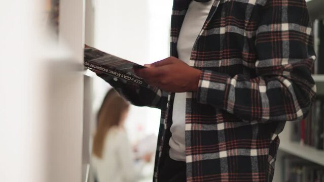 Black man holds book in university library closeup. Engrossed reader reads textbook standing near rack in bookshop. Student chooses scientific literature