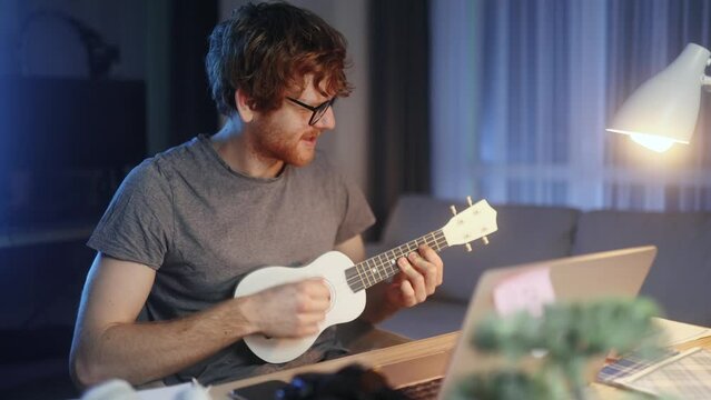 Handsome young red haired man watch video tutorial on ukulele guitar playing or having personal lesson with teacher distance remote on laptop computer late at home Online musical education concept 