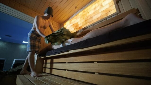 Man lying on wooden bench in SPA with masseur doing massage with oak broom. Client in Finn sauna indoors with professional.