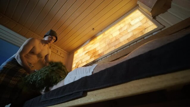 Man lying on wooden bench in SPA with masseur doing massage with oak broom. Client in Finn sauna indoors with professional.