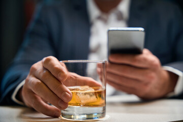 Focus on glass cup with cognac in hands of blurred businessman using smartphone