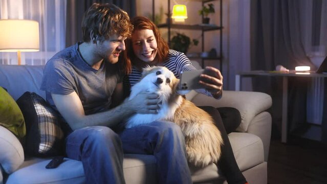 Portrait of young red hair couple with corgi dog pet grimacing taking selfie photograph saving great memories on smartphone mobile phone at home at night Happy relationship concept