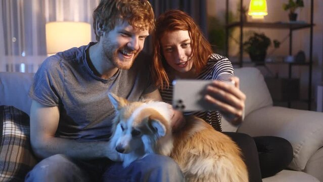 Portrait of young red hair couple with corgi dog pet grimacing taking selfie photograph saving great memories on smartphone mobile phone at home at night Happy relationship concept