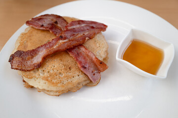 Two crispy bacon strips on a pile of pancakes. There is also a pot of golden maple syrup on the white plate. - 713567809