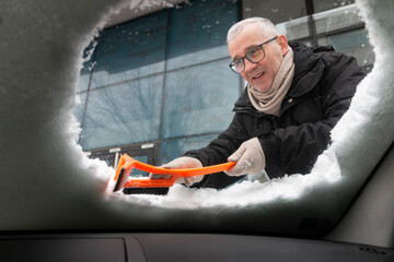 A mature man cleans his car from snow on a frosty winter day. A man scratches the front window of his car on a cold winter morning, interior view