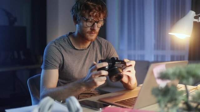 Handsome red haired young man photographer hold digital camera looking at screen choosing photos for editing sitting in front of computer at home workplace at late evening