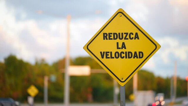 Close up of yellow rhombus traffic road sign reduce speed ahead in spanish. Many cars passing by the asphalt road in the blurred background. Driving rules in South America