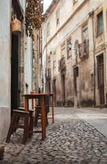 Charm of a cloudy Coimbra morning, rustic wooden tables line a narrow, uphill stone street. A bottle of beer and cup rest on tables with wooden stools, set against building adorned with wall lanterns.