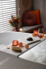 White wooden tray with glass of rose wine, book and burning candles on bathtub in bathroom