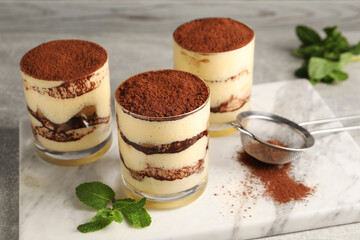 Delicious tiramisu in glasses, mint and sieve with cocoa powder on table, closeup