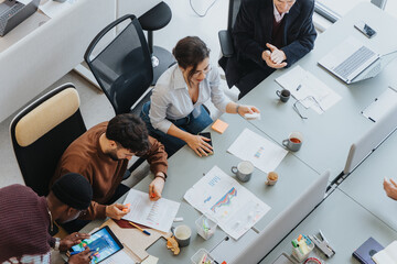 A top view of a multicultural group of professionals collaborating in a business meeting around a table filled with charts, digital devices, and notes. Concept of teamwork and strategy planning.