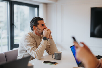 Pensive male in a casual sweater sits in a bright office setting, contemplating during a meeting with a colleague using a smart phone.
