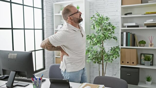 Unhappy young man worker suffering from backache while standing indoors at the office, serious business professional touching injured spinal column, worried about painful back injury at workplace