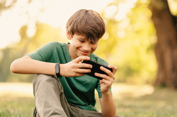 Cheerful teen boy is playing on his phone after classes sitting in a park in autumn.