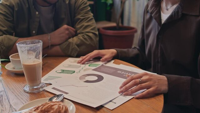 Close-up shot of hands and midsection of unrecognizable woman sitting at table in cafe or coworking, demonstrating business graphs and sales analysis, and presenting marketing strategy to male partner