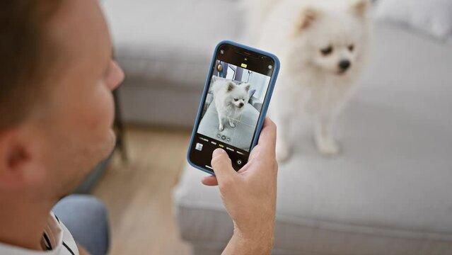 Young caucasian man sitting on the floor at home, relaxed and focused while making a photo of his adorable dog using smartphone
