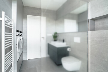 A small bathroom with a toilet, sink, washing machine and dryer. The walls and the floor are made of gray tiles. There is a big milky glass wall, which separates shower from the rest of the bathroom.