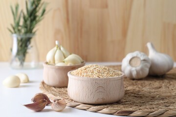 Dehydrated garlic granules in bowl, fresh bulbs and cloves on white table