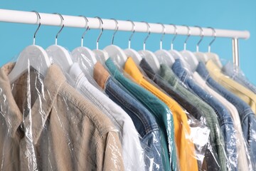 Dry-cleaning service. Many different clothes in plastic bags hanging on rack against light blue background, closeup