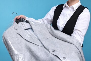 Dry-cleaning service. Woman holding jacket in plastic bag on light blue background, closeup