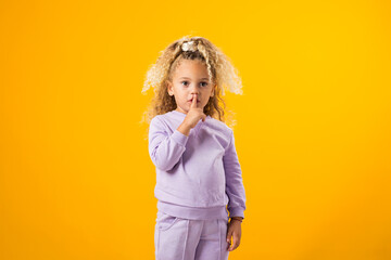 Child girl touching her lips with finger, demonstrate shh gesture isolated on yellow background