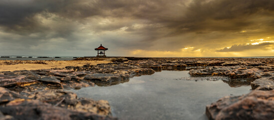 A picturesque gazebo temple stands atop the rocky beach, framed by billowing clouds and a vibrant...