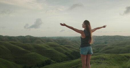 Woman stand on green hill meadow, raised hands, wind blow her hair - freedom, enjoy nature. Tourist...