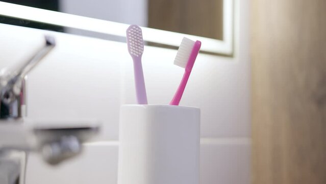 close-up shot woman hand placing purple toothbrush back into glass on edge of sink in white-tiled bathroom. image represents concept of brushing teeth in morning using purple brush.