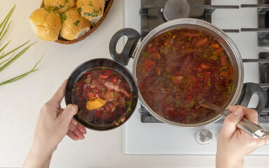 Women's hands pour Ukrainian borsch from a pot into a plate, top view.