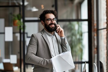 Smiling happy young bearded Latin professional business man executive holding documents and cell phone making mobile call at work on cellphone consulting client standing in modern office