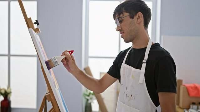 Young, handsome hispanic man engrossed in drawing, inside the vibrant art studio, the talented artist stands in apron, paintbrush in hand, studiously concentrating on his canvas