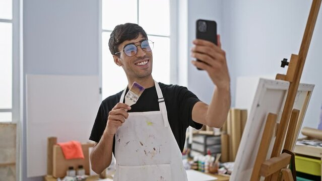 Smiling young hispanic man artist taking a splendid selfie picture with his smartphone at the bustling art studio, canvas in one hand, paintbrush in the other!